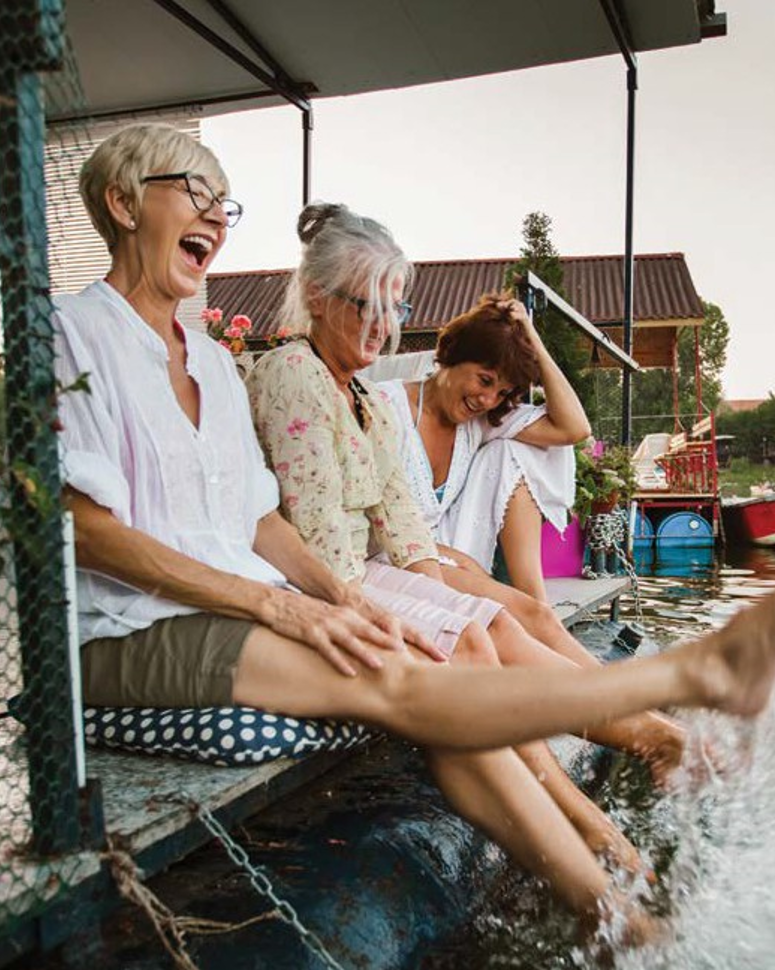 Three women sit along a dock while laughing